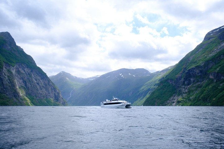 Ferry on a fjord with steep, green mountains under a cloudy sky.