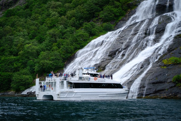 Tour boat near a waterfall cascading down a rocky hillside surrounded by lush greenery.