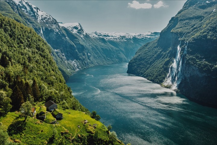 Scenic view of a fjord with lush greenery, snowy peaks, and a waterfall cascading down a cliffside.